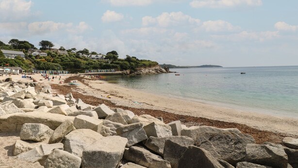 A coastal beach with large rocks in the foreground, a sandy shore, and houses on a hillside in the background. - Manuka (Falmouth)