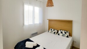 A neatly made bed with a dark blue blanket and two rolled towels, a wooden headboard, a woven pendant light, and a window with white curtains. - Tourné Vers le sud (Linguizzetta)