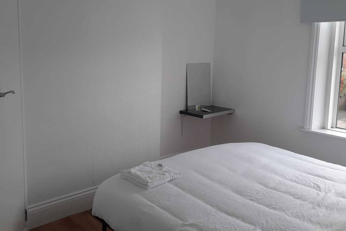 Een slaapkamer met een bed, nachtkastje, spiegel en raam.