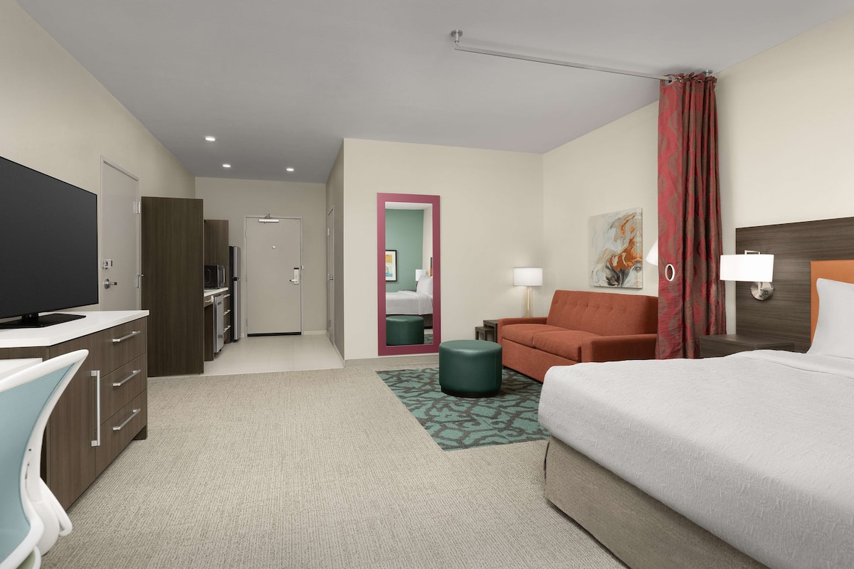 Een hotelkamer met een bed, een bureau met een televisie, een bank, een klein voetenbankje en een badkamer zichtbaar door een geopende deur.