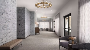 A modern hotel hallway with patterned walls, a chandelier, seating area, and a view through a glass door.