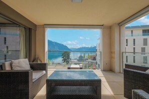 Un balcone con mobili in vimini, un tavolino in vetro e vista sulle montagne e su un lago.