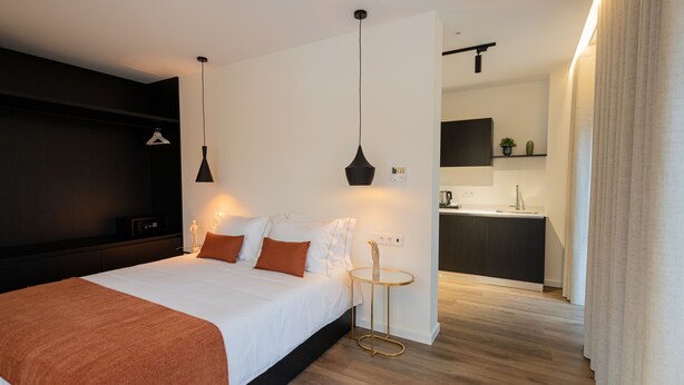 A modern hotel room with a large bed, bedside tables, a small round table, a pendant light, a wall-mounted shelf, and a view into the kitchen area. - Watts House (Seia)