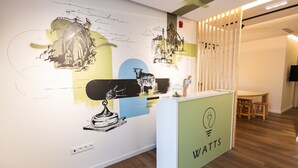 A reception desk with a logo, a wall with artwork, and a wooden partition. - Watts House (Seia)