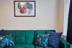 Un sofá verde con cojines estampados, una obra de arte abstracta enmarcada en la pared y una ventana con cortinas.
