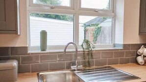 A kitchen with a stainless steel sink, a window with a view of a house, and a potted plant on the windowsill. - Elfleda Stay | Modern 5 bedroom house (Cambridge)