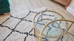A room with a white rug featuring a black geometric pattern, a round glass table with a gold frame, and a wooden cabinet in the background. - Elfleda Stay | Modern 5 bedroom house (Cambridge)