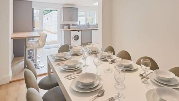 A dining area with a long table set for six, chairs, and a kitchen visible in the background. - Elfleda Stay | Modern 5 bedroom house (Cambridge)