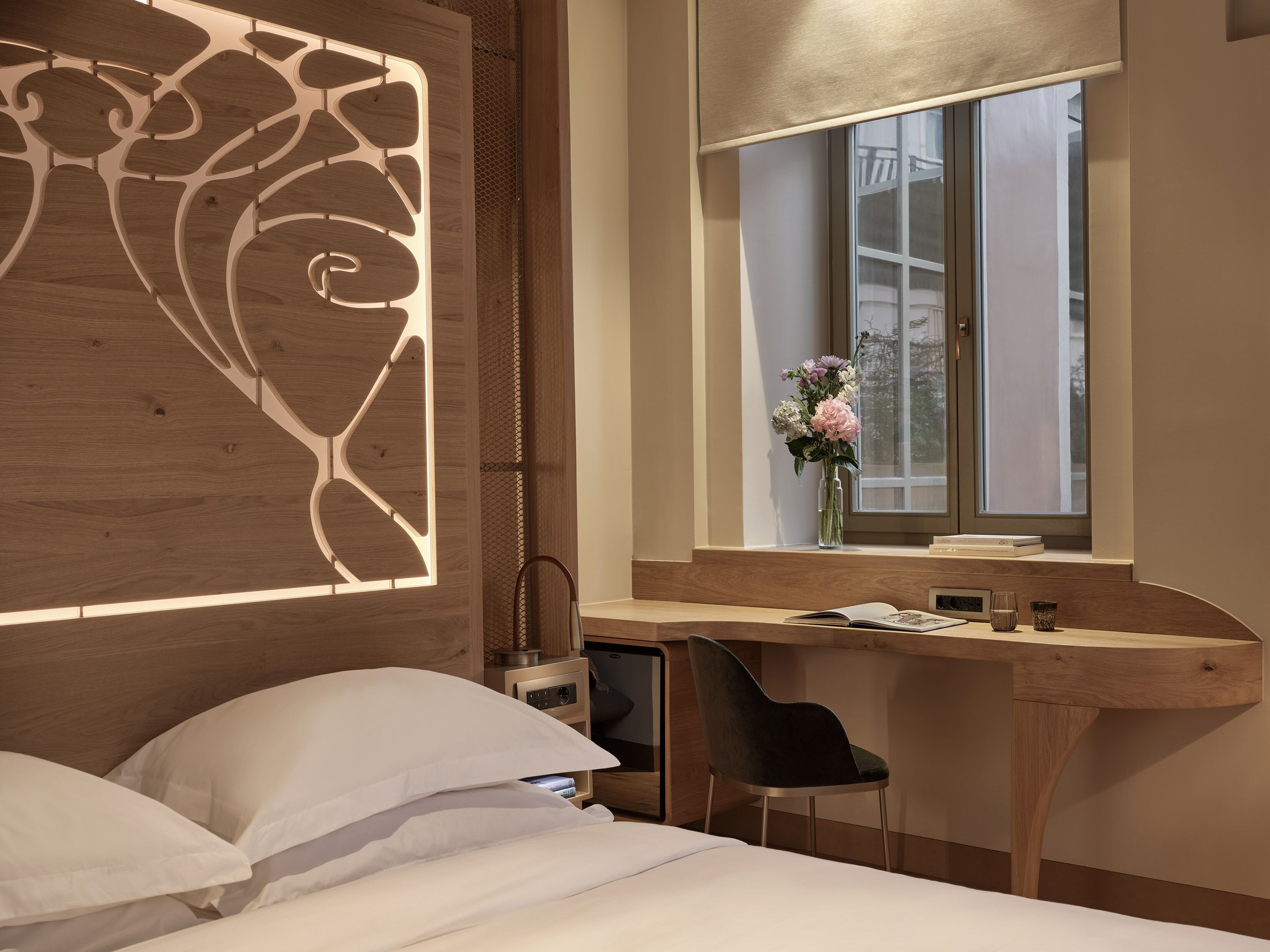 A modern bedroom with a wooden headboard, a bedside table with a vase of flowers, a desk with a chair, and a large window.