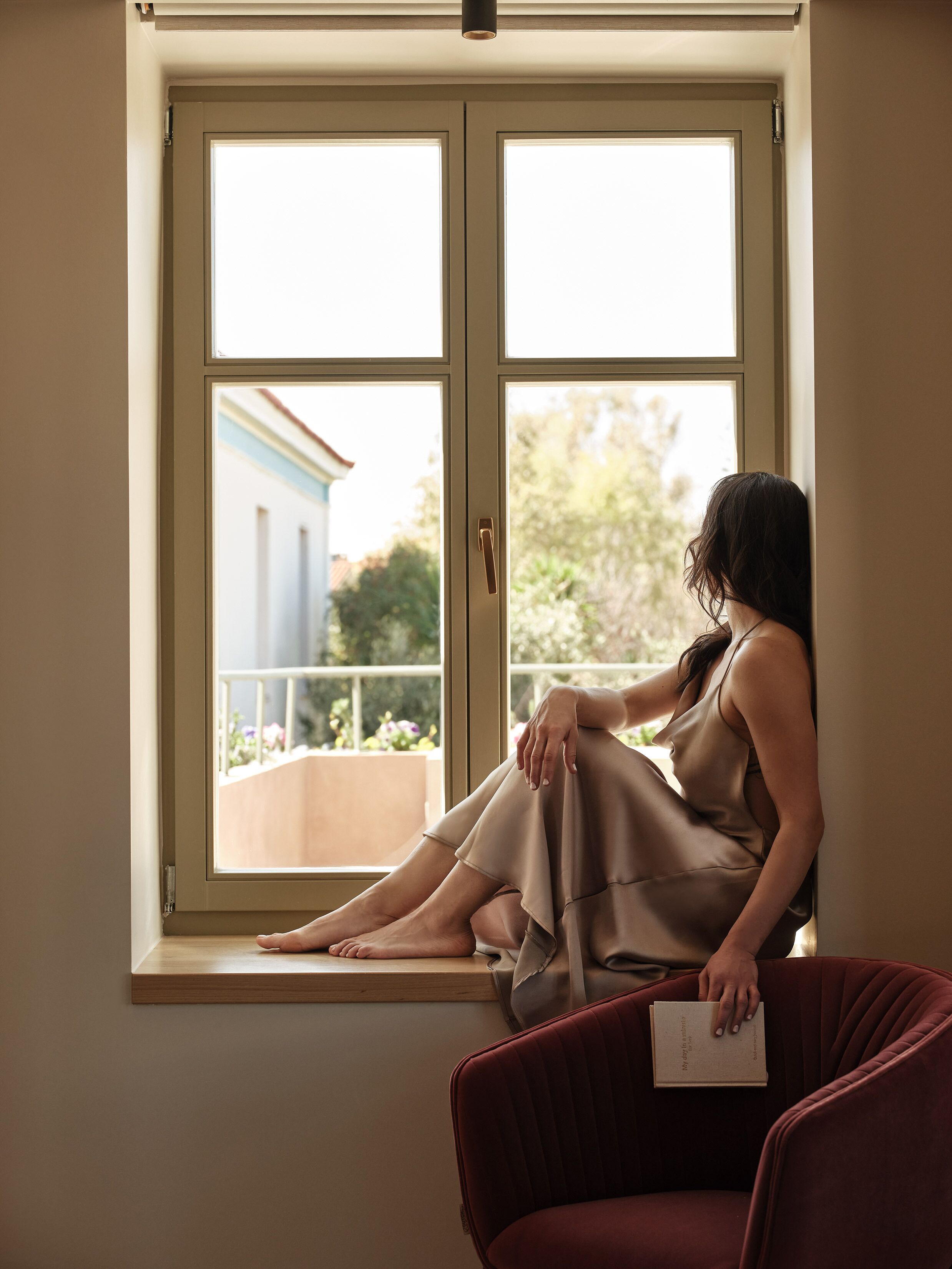 A woman sitting by a window with a view of a balcony and greenery.