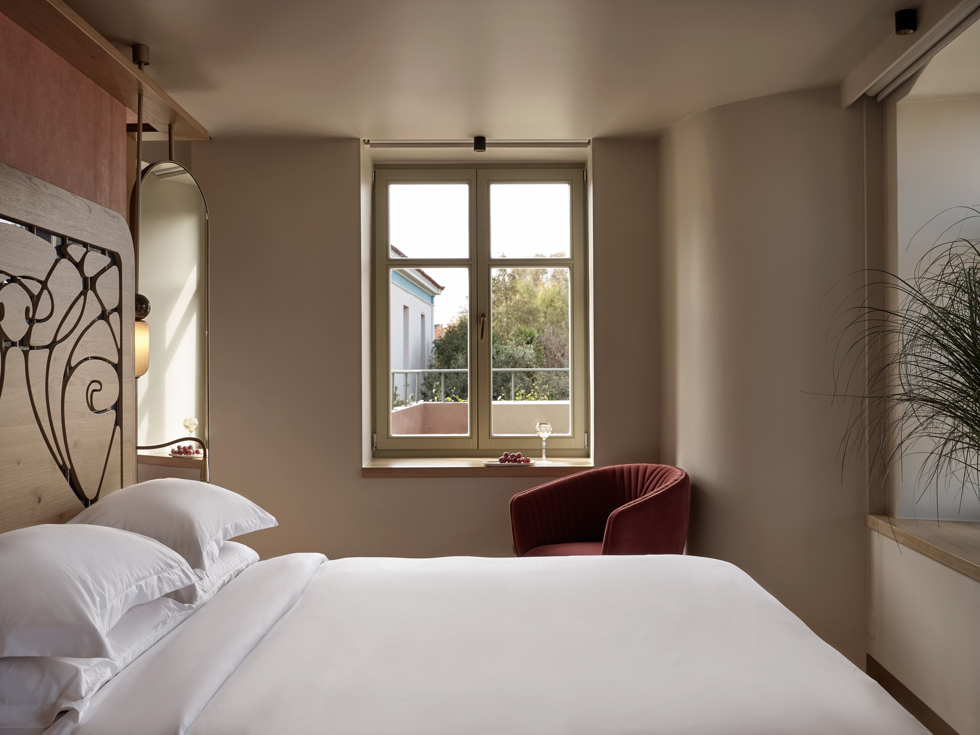 A bedroom with a large bed, a window with a view of the outside, a red chair, and a plant.