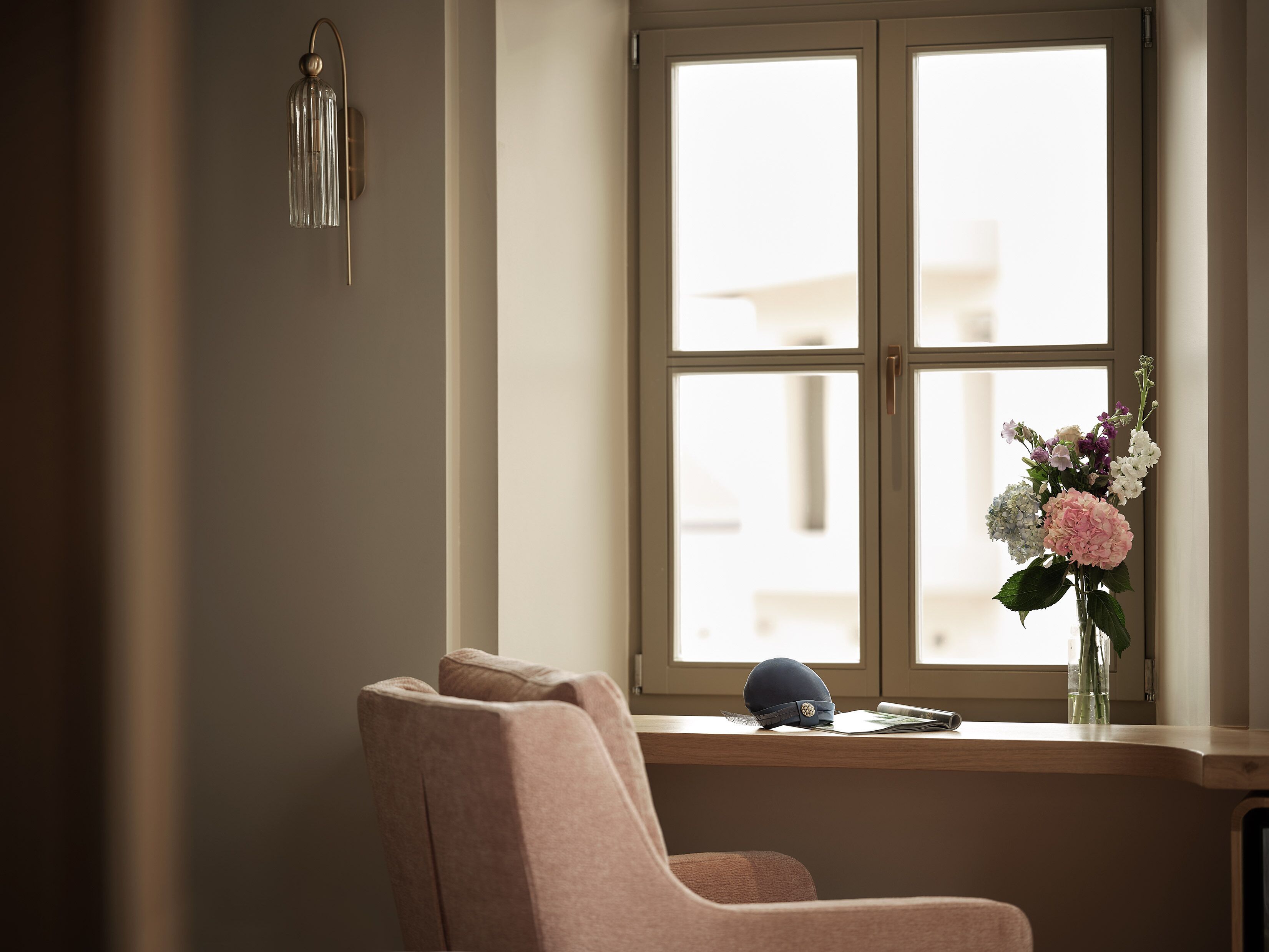 A room with a window, a chair, a vase with flowers, and a wall-mounted light fixture.