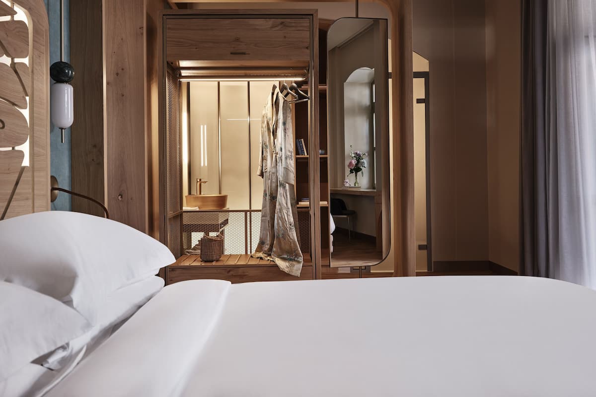 A modern hotel room with a wooden interior, a large bed, a bathroom with a wooden basin, and a view into the adjacent room.