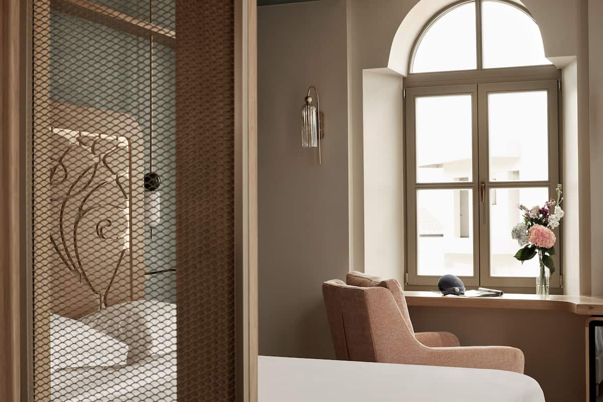 A bedroom with a large window, a chair, a bedside table, and a decorative wall piece.