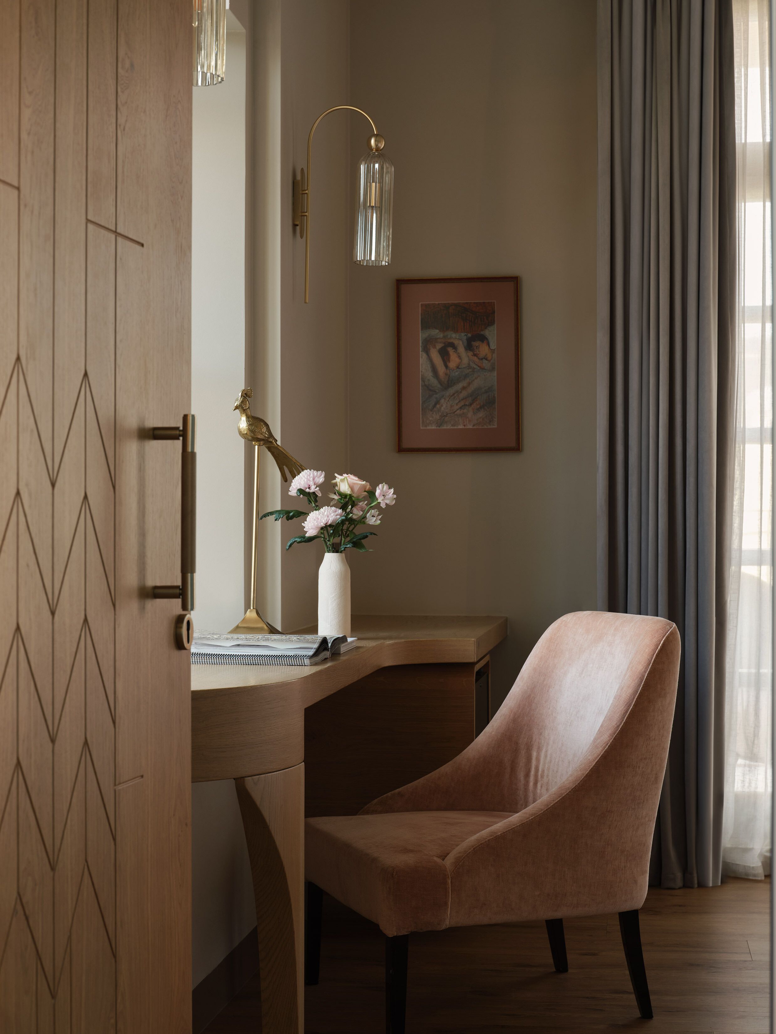 Una habitación moderna con un escritorio de madera, una silla rosa, un florero con flores, un cuadro enmarcado y una lámpara.