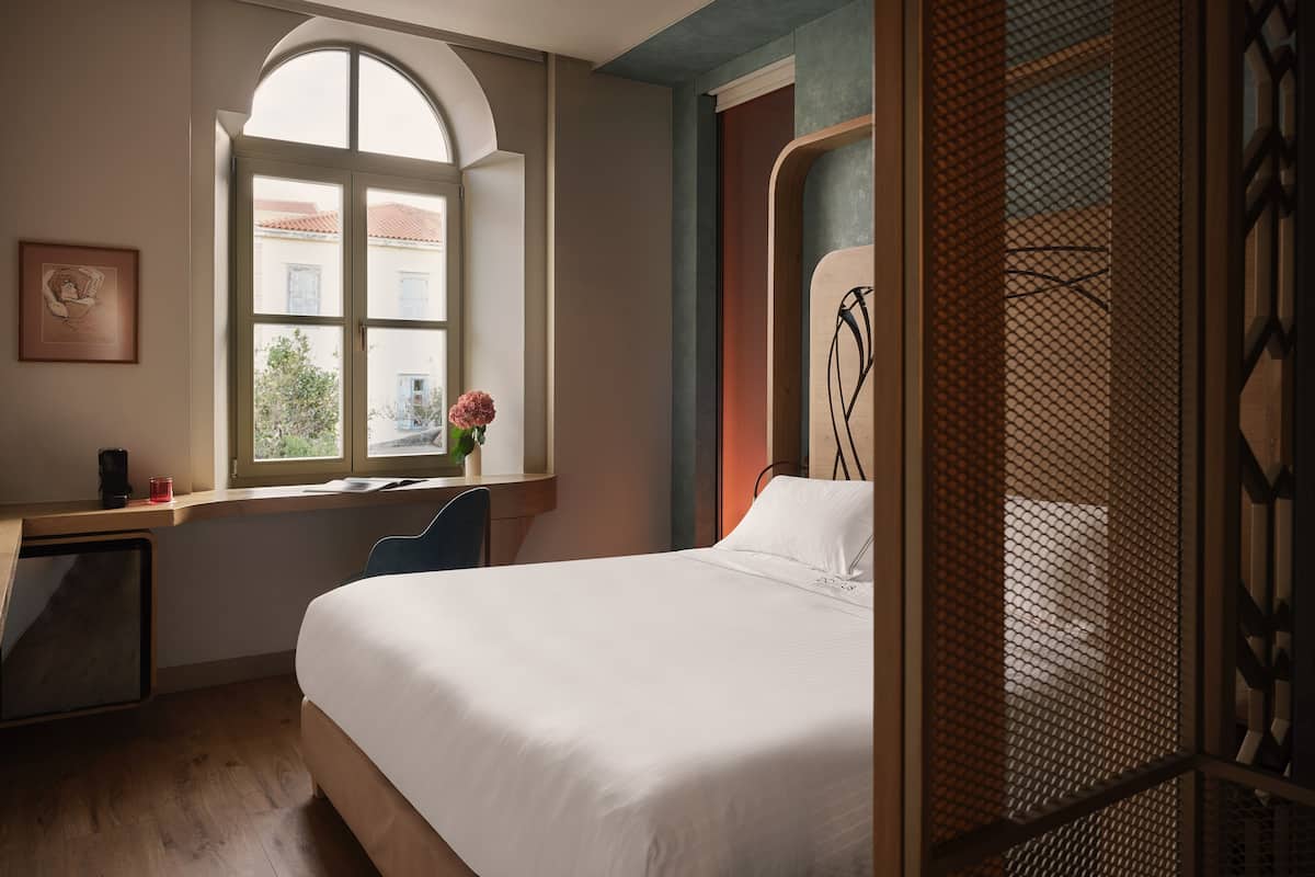 A bedroom with a large bed, a desk with a chair, a window with a view of buildings, and a decorative wall panel.