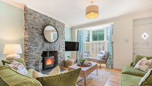 Ein gemütliches Wohnzimmer mit Kamin aus Stein, grünen Sofas, einem Flachbildfernseher und einem großen Fenster mit Vorhängen.