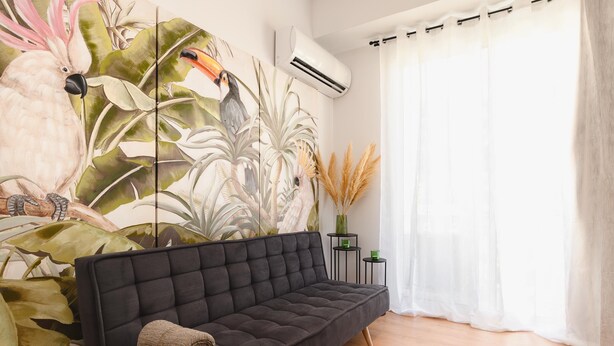 Una sala de estar moderna con un sofá oscuro, un mural tropical grande, una mesita auxiliar con un florero y una ventana con cortinas translúcidas.