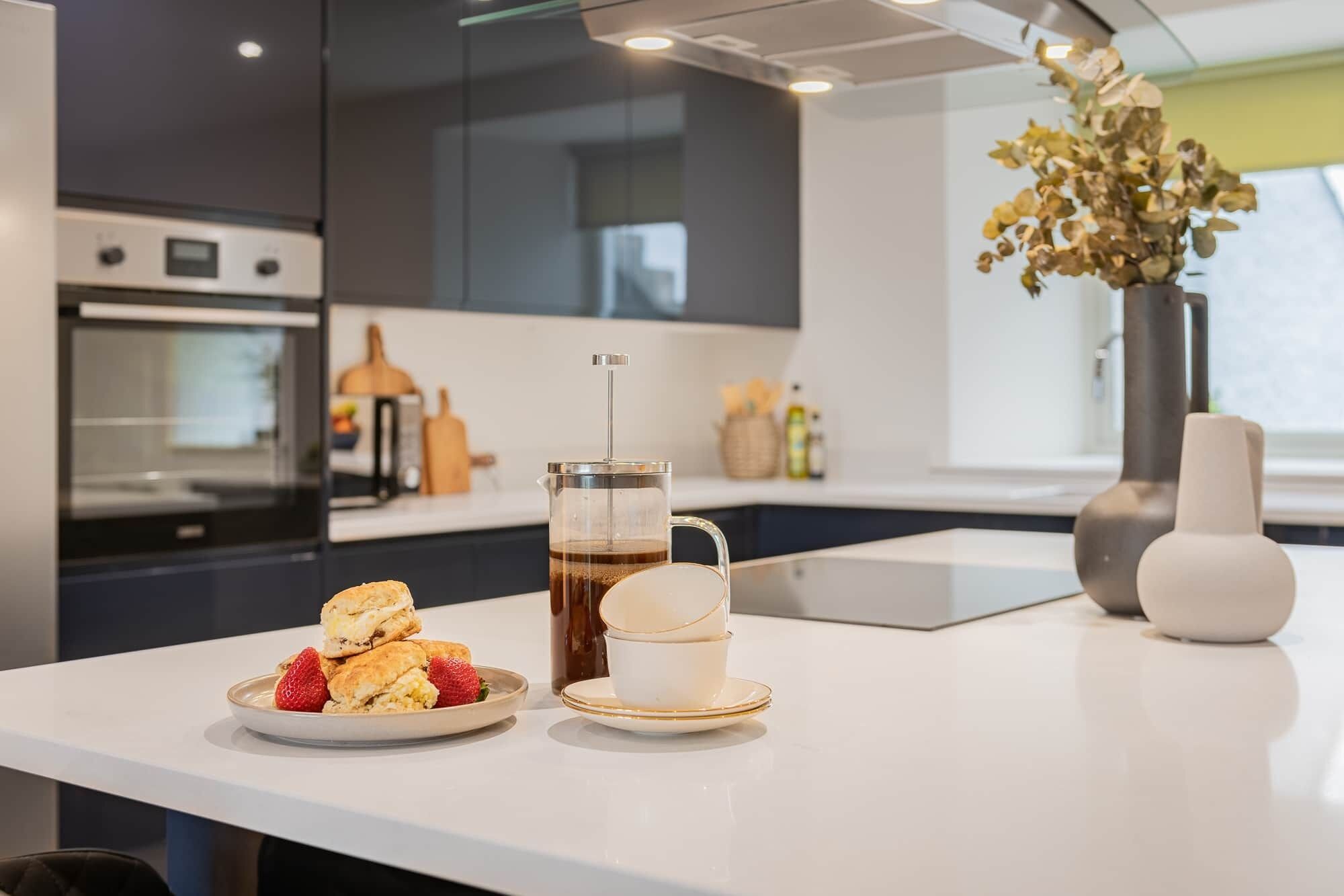 A modern kitchen with a coffee maker, a plate of scones, and a cup of coffee on a white countertop.