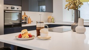 Une cuisine moderne avec une machine à café, une assiette de scones et une tasse de café posée sur un plan de travail blanc.