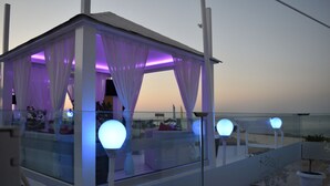 A glass-enclosed outdoor seating area with white curtains and illuminated spherical lights, overlooking a body of water. - Pola Costa Beach Hotel Apartment (Paralimni)