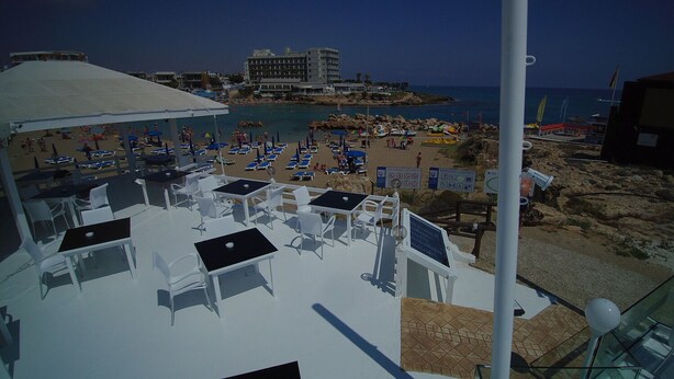 A beachside terrace with white tables and chairs, a covered area, and a view of the beach and ocean. - Pola Costa Beach Hotel Apartment (Paralimni)