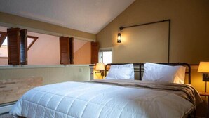 A bedroom with a large bed, wooden shutters, a wall-mounted lamp, and a window with shutters. - Cozy Kettlebrook 2BR: Ski Access, Fireplace & Deck (Ludlow)