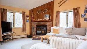 A living room with a fireplace, a sofa, a television, and a large window. - Cozy Kettlebrook 2BR: Ski Access, Fireplace & Deck (Ludlow)