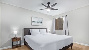 Ein Schlafzimmer mit einem Bett, einem Nachttisch, einem Deckenventilator, einem Fenster mit Vorhängen und einem Bild an der Wand.