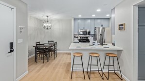 Eine moderne Küche mit grauen Schränken, einer weißen Arbeitsplatte und einem Edelstahl-Kühlschrank. Drei Barhocker stehen an der Theke, und über dem Esstisch hängt eine Pendelleuchte.