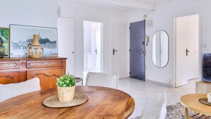 Een moderne woonkamer met een houten eettafel, een bank en een televisie op een dressoir. Er zijn twee deuren, waarvan één naar een badkamer en de andere naar een slaapkamer.