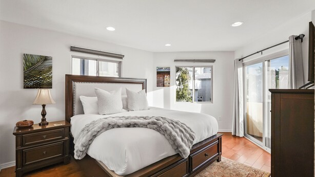A bedroom with a large bed, a nightstand with a lamp, and a view of the outdoors through a sliding door. - Pacific Beach Gem: Rooftop Deck, Bay Views, AC & Pet Friendly (San Diego)