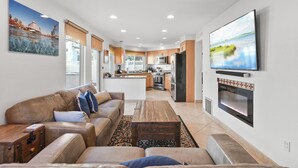 A modern living room with a large TV, leather sofas, and a kitchen in the background. - Pacific Beach Gem: Rooftop Deck, Bay Views, AC & Pet Friendly (San Diego)