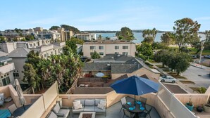 A rooftop patio with outdoor furniture, a blue umbrella, and a view of the city and water. - Sail Bay Dream | Rooftop Patio, Bay Views, AC, Garage, Pinball, Paddle Boards + Bikes (San Diego)