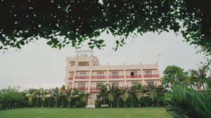 A multi-story building with a flat roof, surrounded by a well-maintained lawn and various tropical plants. - Kashi Anandam Spiritual & Wellness Vedic Village (Varanasi)