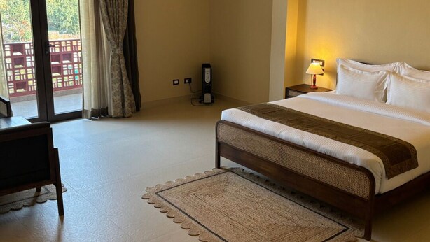 A hotel room with a large bed, a desk, a chair, a bedside table with a lamp, and a view through a sliding door. - Kashi Anandam Spiritual & Wellness Vedic Village (Varanasi)