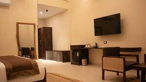 A hotel room with a bed, a TV mounted on the wall, a desk with a chair, and a small kitchenette area. - Kashi Anandam Spiritual & Wellness Vedic Village (Varanasi)