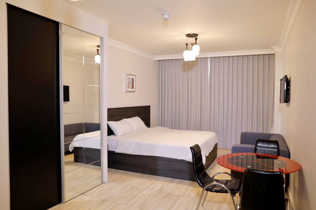 A modern hotel room with a large bed, a glass table, a sofa, and a mirror.