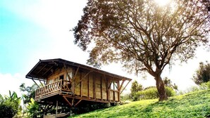 A wooden structure with a balcony, situated on a grassy slope with a large tree nearby. - Casa Laureles (Filandia)
