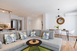 Un salon moderne avec un canapé d’angle gris, une table basse ronde et un coin repas comprenant une table et des chaises blanches.