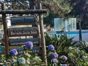 A wooden sign with text in a foreign language, a pool, and some purple flowers. - Pousada Xodó das Montanhas (Afonso Claudio)