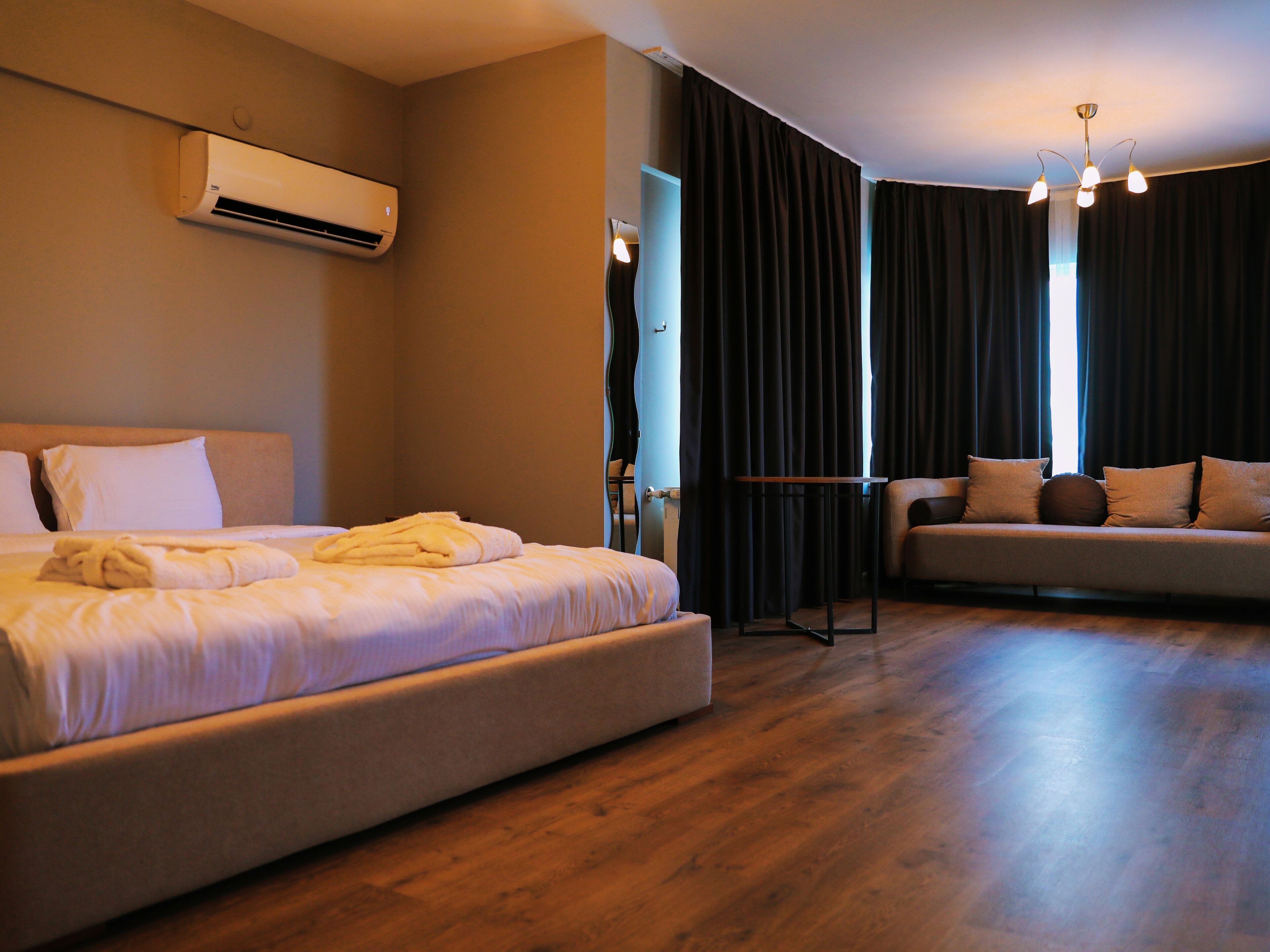 A hotel room with a large bed, a sofa, a small table, and a ceiling-mounted air conditioner.