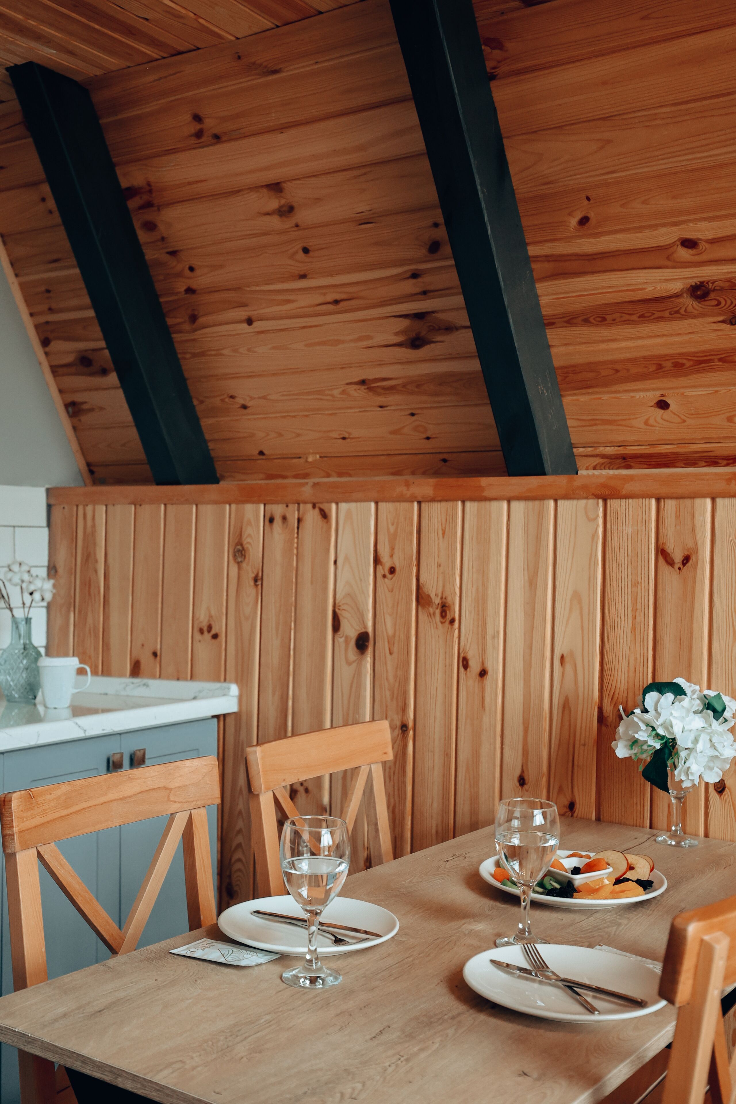 Una mesa de comedor de madera dispuesta con platos, cubiertos y vasos, con una pared y techo de madera al fondo.