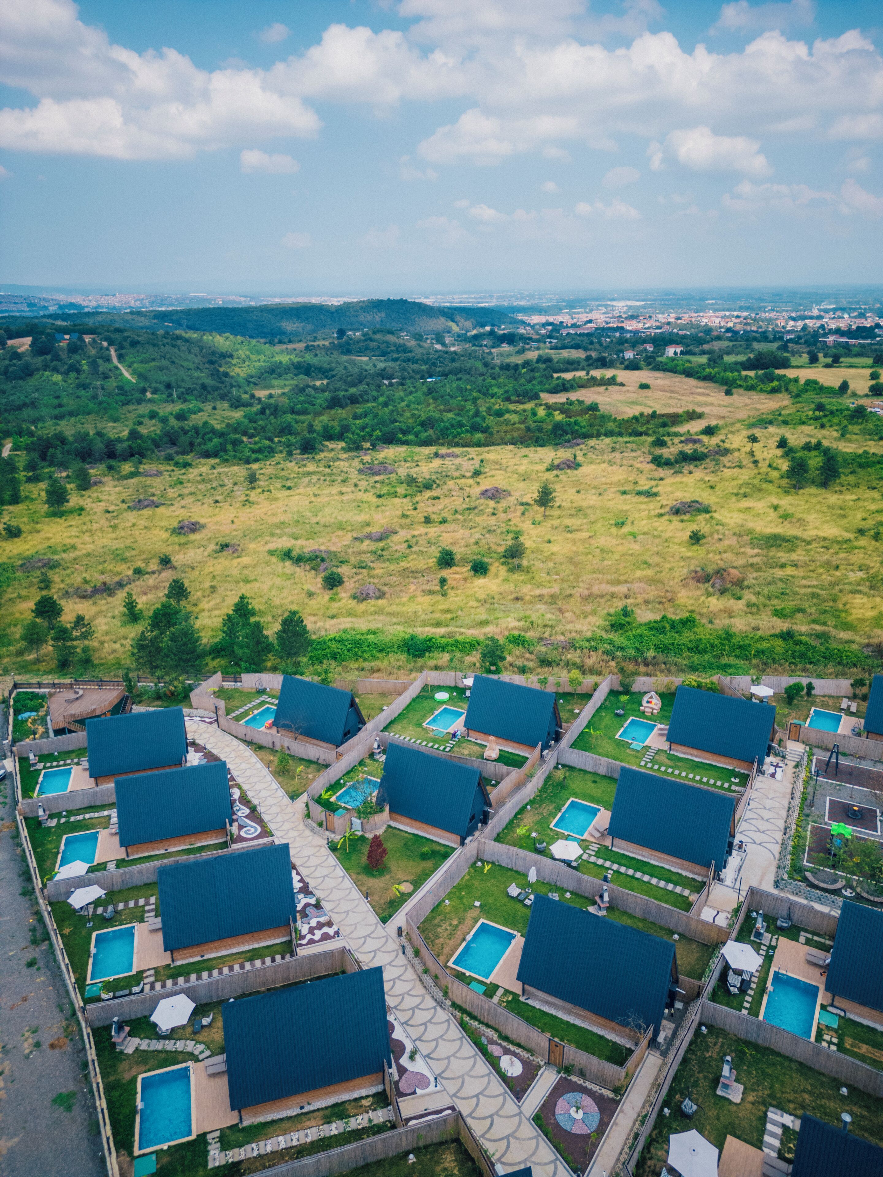 An aerial view of a resort with multiple swimming pools, each surrounded by a covered deck area. The resort is set in a green landscape with a distant view of a city skyline.