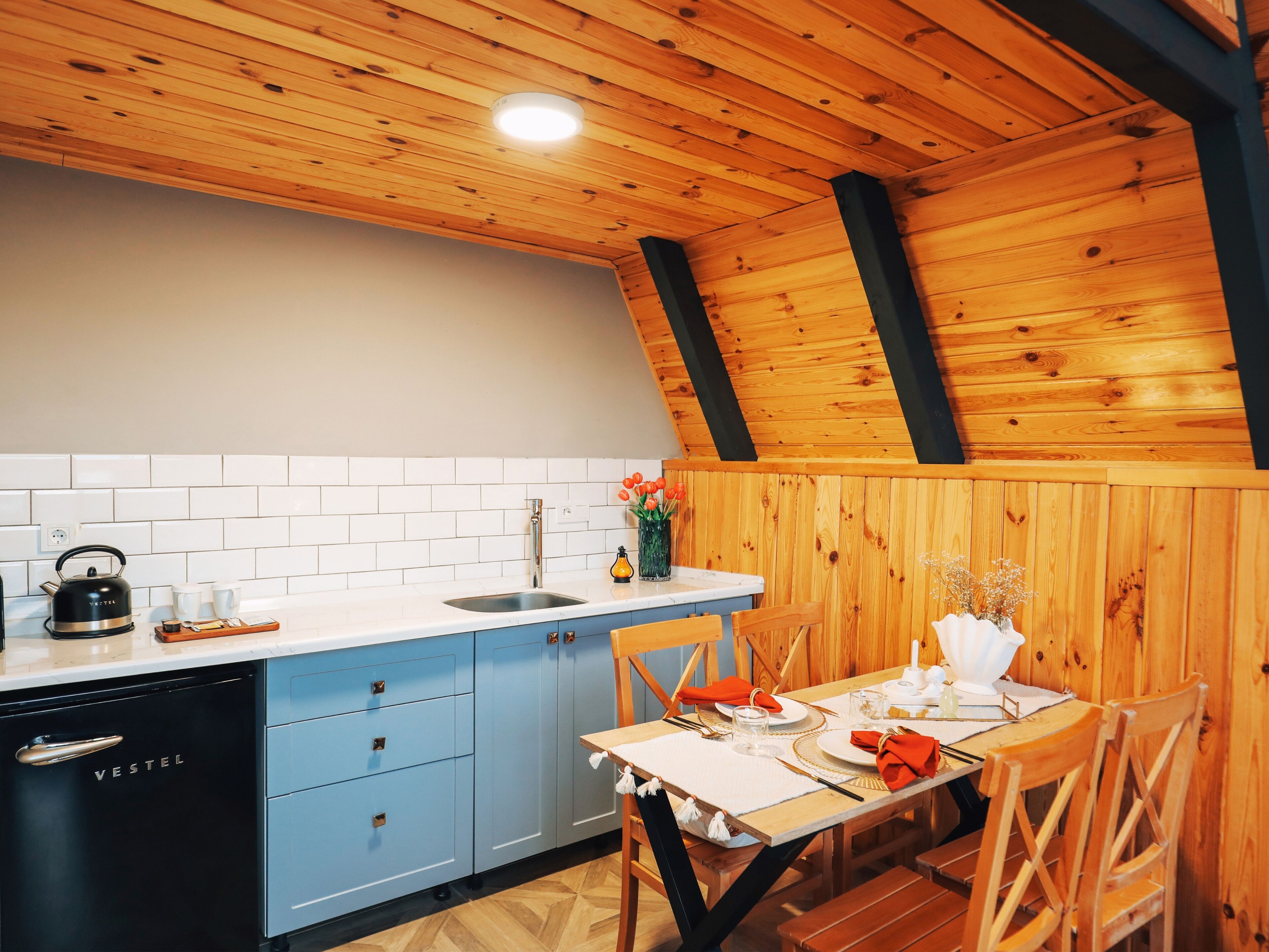 A kitchen with a wooden ceiling, white tiled backsplash, blue cabinets, and a dining area with a table set for two.