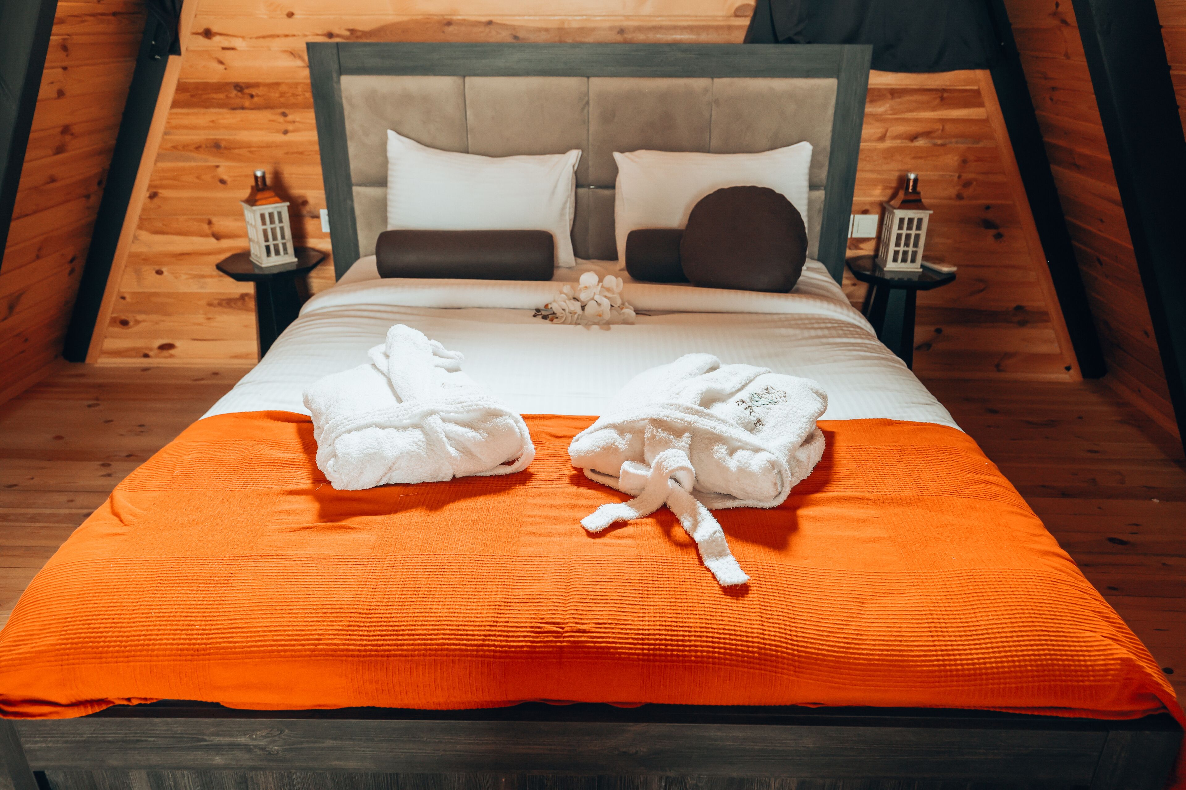 Een net opgemaakt bed met een wit-oranje deken, twee witte handdoeken en een hoofdbord met twee witte en twee bruine kussens.