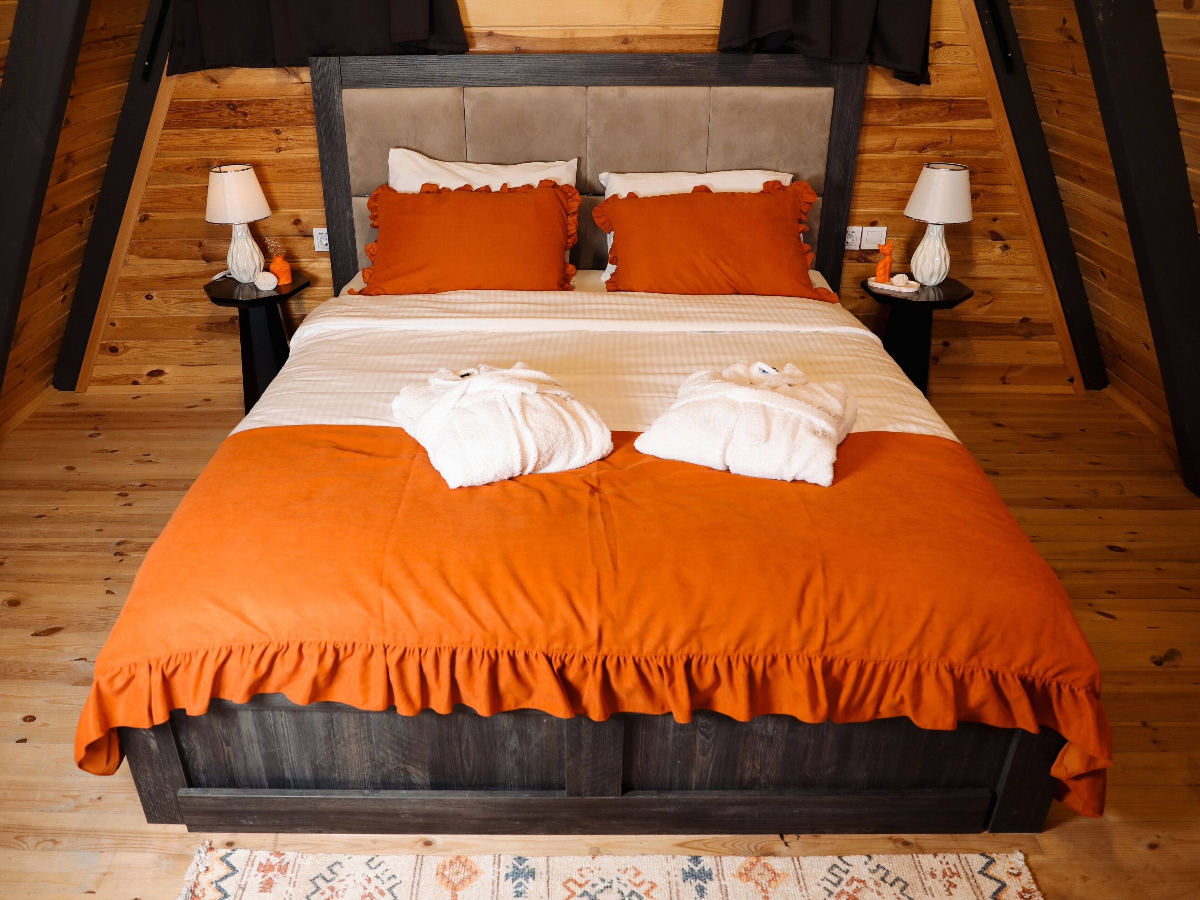 Un lit bien fait, avec une couvre-lit et des oreillers orange, deux serviettes blanches et deux lampes de chevet posées sur des tables de nuit en bois.
