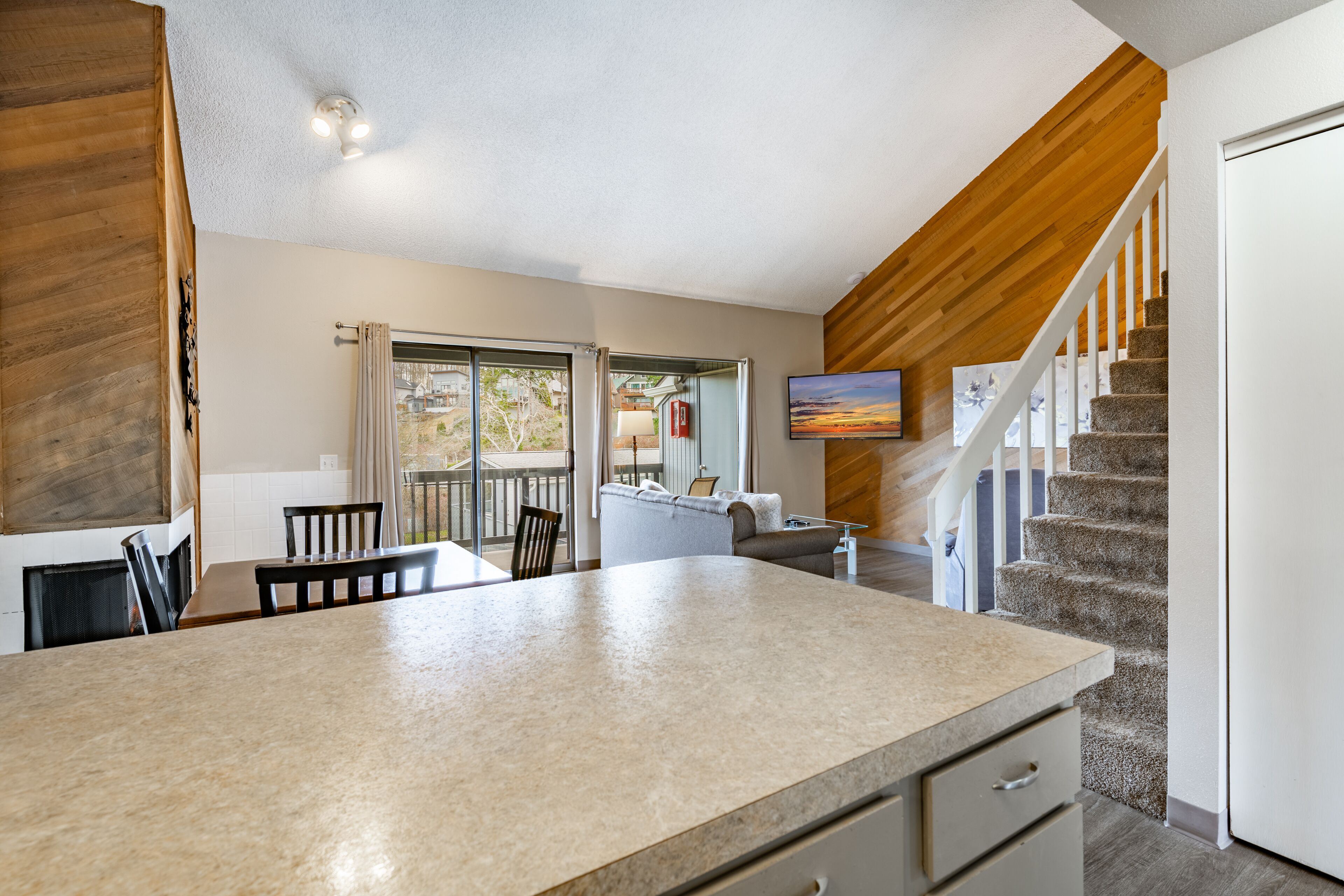 A modern kitchen with a large island, a dining area with chairs, a staircase, and a living area with a TV.