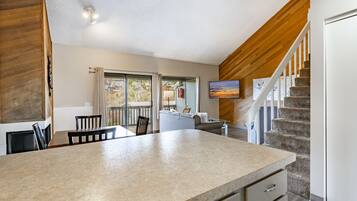 A modern kitchen with a large island, a dining area with chairs, a staircase, and a living area with a TV.