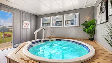 A hot tub in a room with wooden flooring, a view of the backyard, and framed pictures on the wall.
